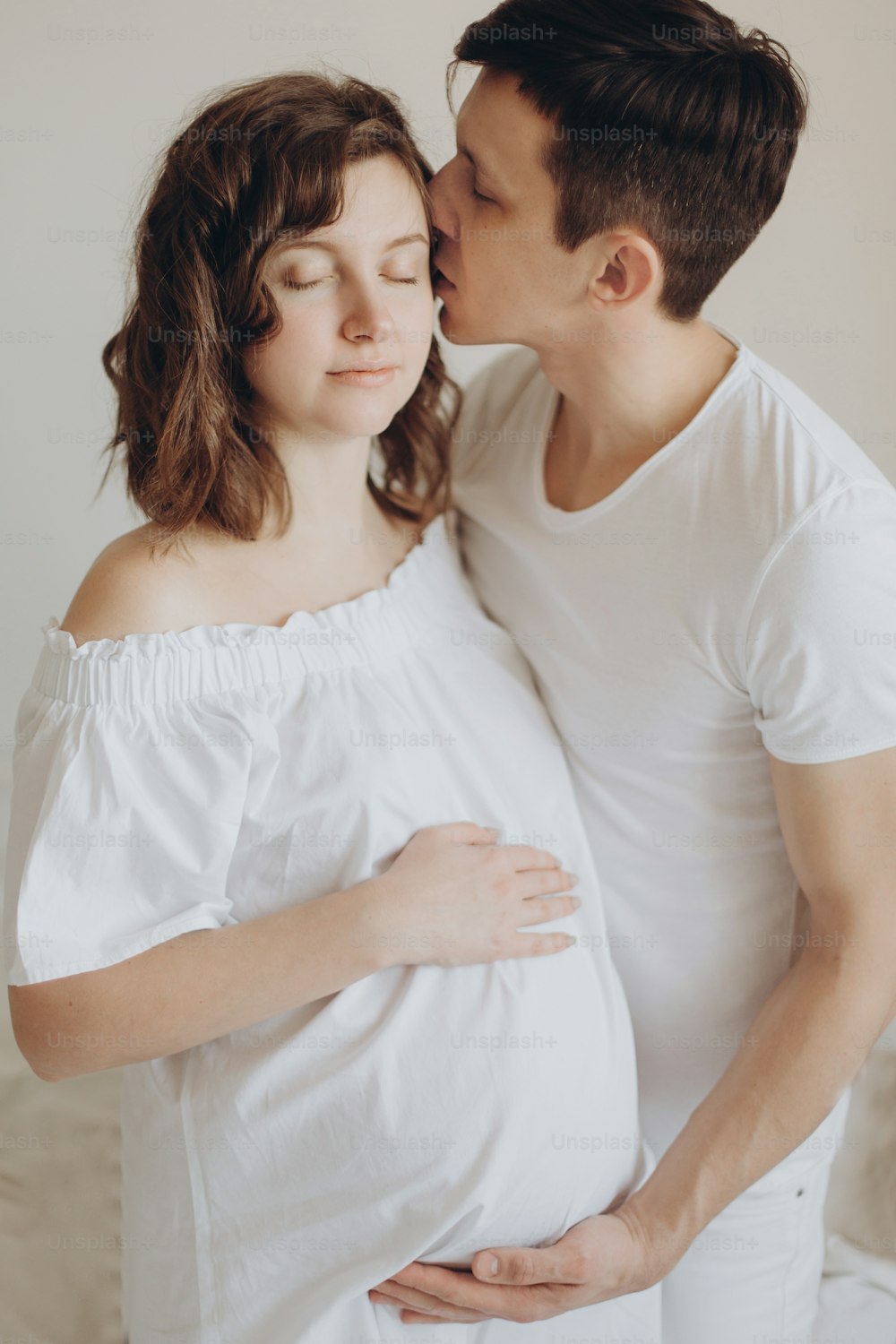 白いベッドでリラックスし、腹の隆起を保持している幸せな妊娠中のカップル。笑顔の妻にキスをし、赤ちゃんのお腹を抱きしめる幸せな若い夫。 自宅で白い服を着たスタイリッシュな妊娠中の家族。本当の幸せ