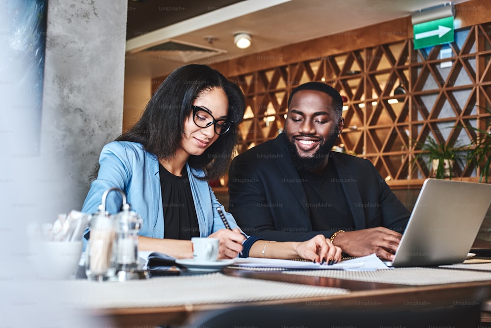 Cheerful businessman and businesswoman having lunch at restaurant, sitting at table near window, drinking coffee, discussing project, smiling cheerful. Man had a look on his colleague, while she is making notes