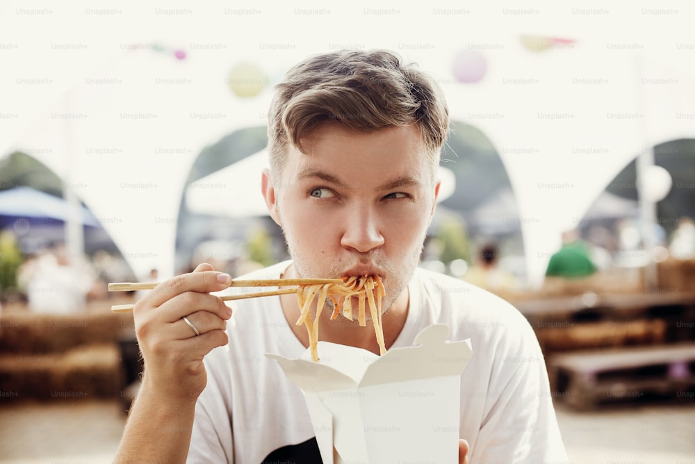 Stilvoller hungriger Mann, der leckere Wok-Nudeln mit lustigen Emotionen aus Karton mit Bambusstäbchen isst. Asiatisches Street Food Festival. Hipster-Verkostungsnudeln in Papierbox zum Mitnehmen