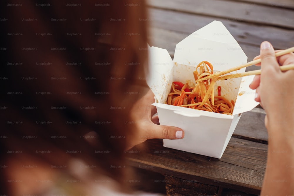 Muchacha comiendo fideos al wok con verduras y mariscos en caja de cartón para llevar, con palillos de bambú, primer plano. Cocina tradicional asiática en el festival de comida callejera. Fideos tailandeses en caja de papel para llevar