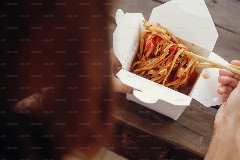 Wok avec nouilles et légumes dans une boîte en carton à emporter et baguettes en bambou. Cuisine asiatique traditionnelle. Festival asiatique de la cuisine de rue. Fille mangeant des nouilles thaïlandaises dans des plats à emporter en boîte ouverte. Livraison de nourriture