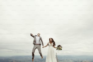 happy gorgeous bride and stylish groom jumping and having fun, boho wedding couple, luxury ceremony at mountains with amazing view, space for text