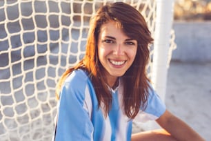Beautiful young female football fan squatting in front of a goal net on a building rooftop, waiting for a match