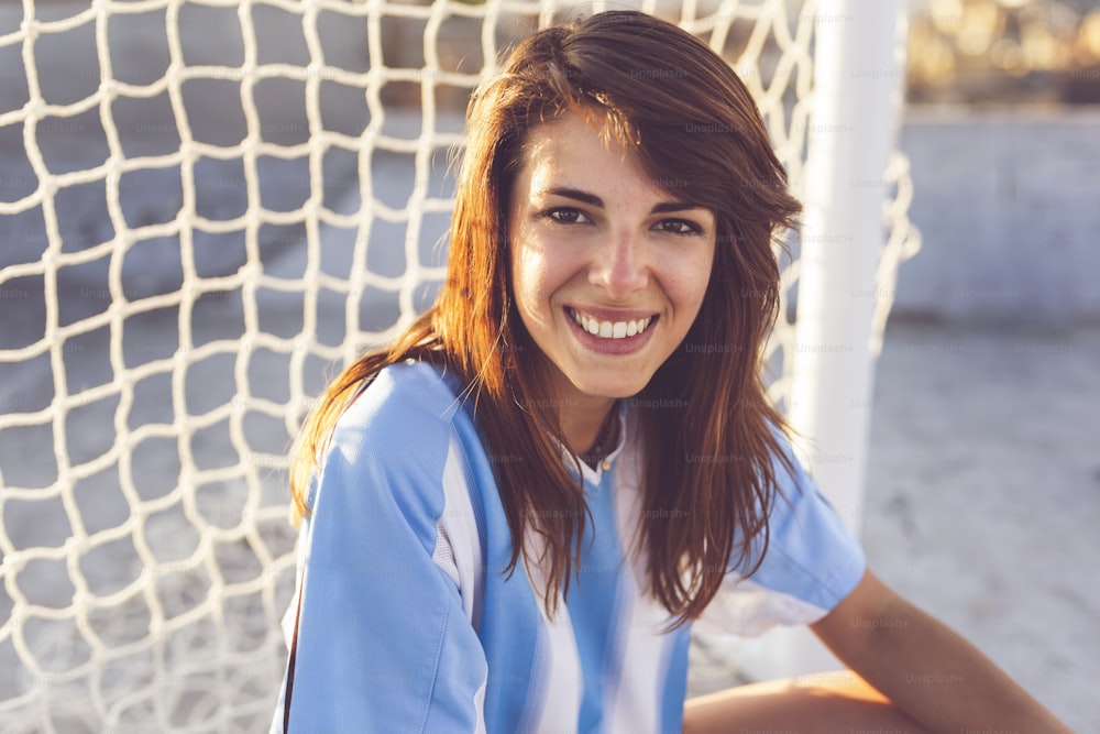Hermosa joven fanática del fútbol en cuclillas frente a una red de gol en la azotea de un edificio, esperando un partido