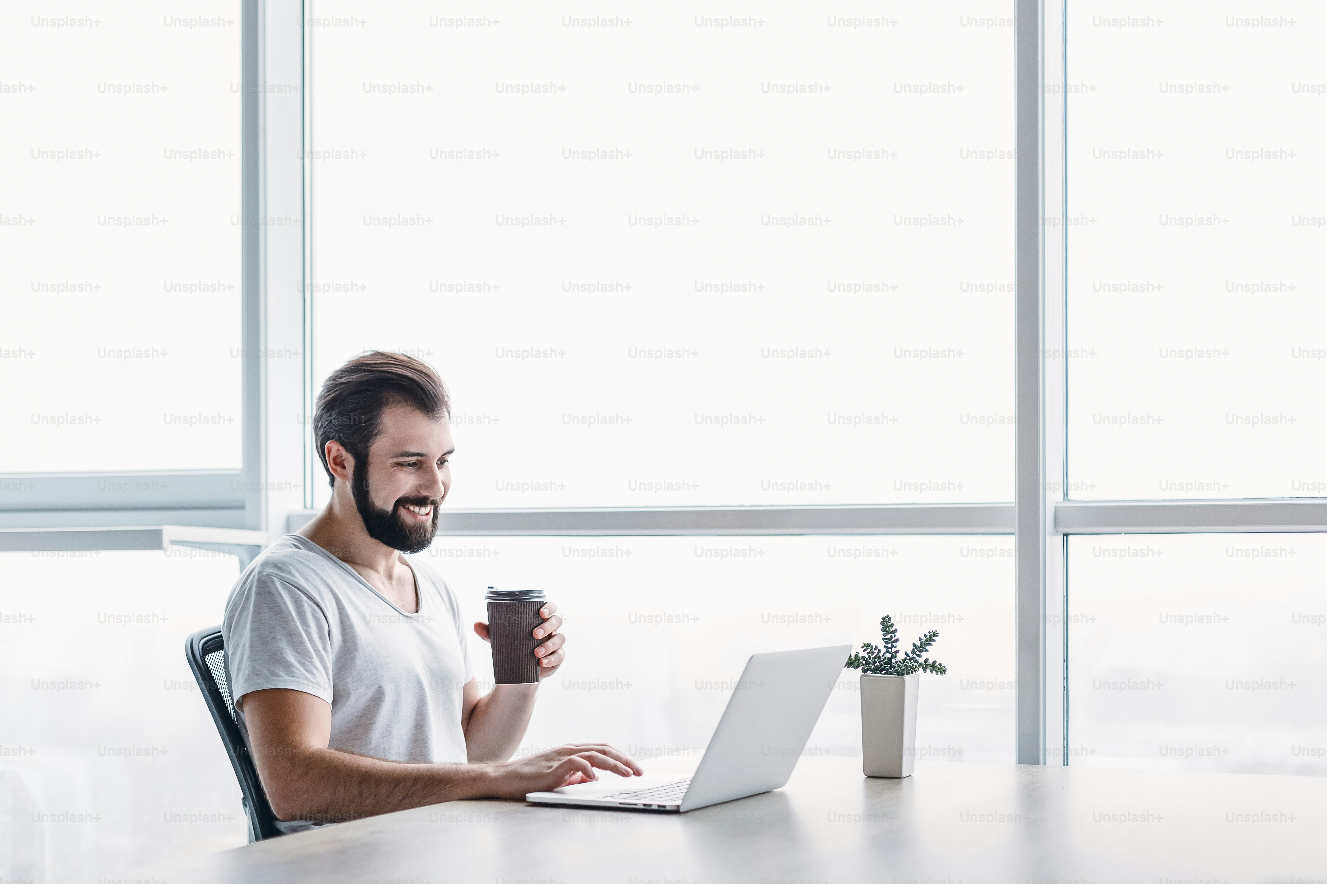 Portrait of handsome happy bearded young businessman in grey T-shirt, sitting in office and have a break with cup of coffee. He uses his laptop, looking on the screen with toothy smile