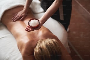 Concept of relaxation and body care. Top angle portrait of therapist holding bowl with mineral salt for making scrubbing and relaxing massage for young lady