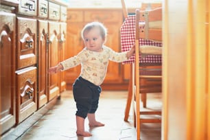 Baby girl standing on the floor in the kitchen and holding on to furniture. Little child oulling up at home