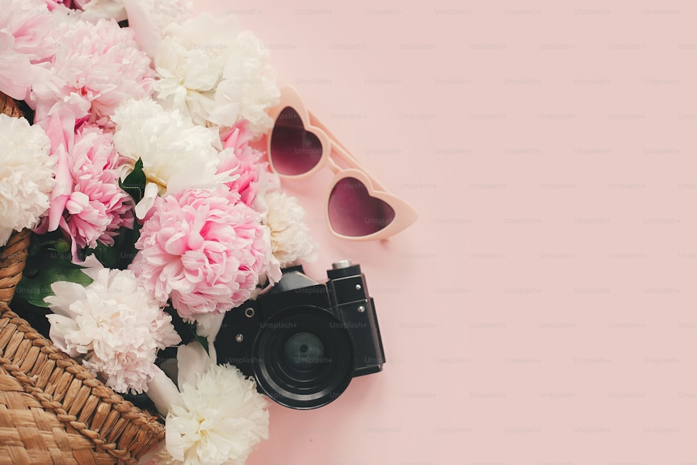 Stylish photo camera, sunglasses, straw  bag with pink and white peonies on pink paper flat lay with space for text. Hello summer.  International womens day.