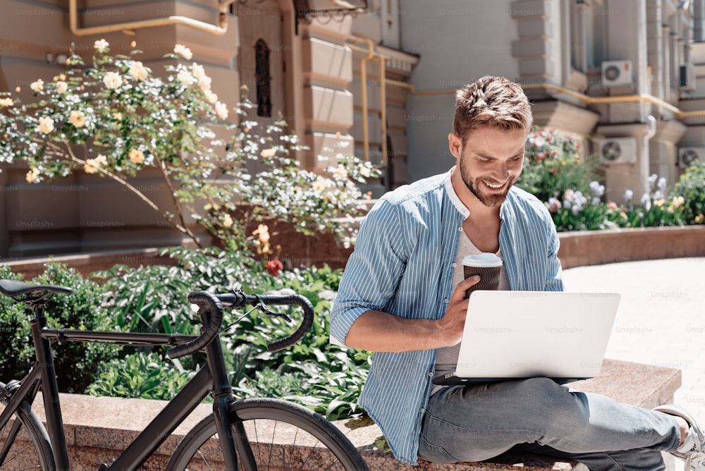 Portrait of handsome young man sitting in the park and holding his laptop and a cup of coffee. Bearded brown-haired man dressed in a blue striped shirt and jeans looking in his laptop and smiling. Summertime, relax