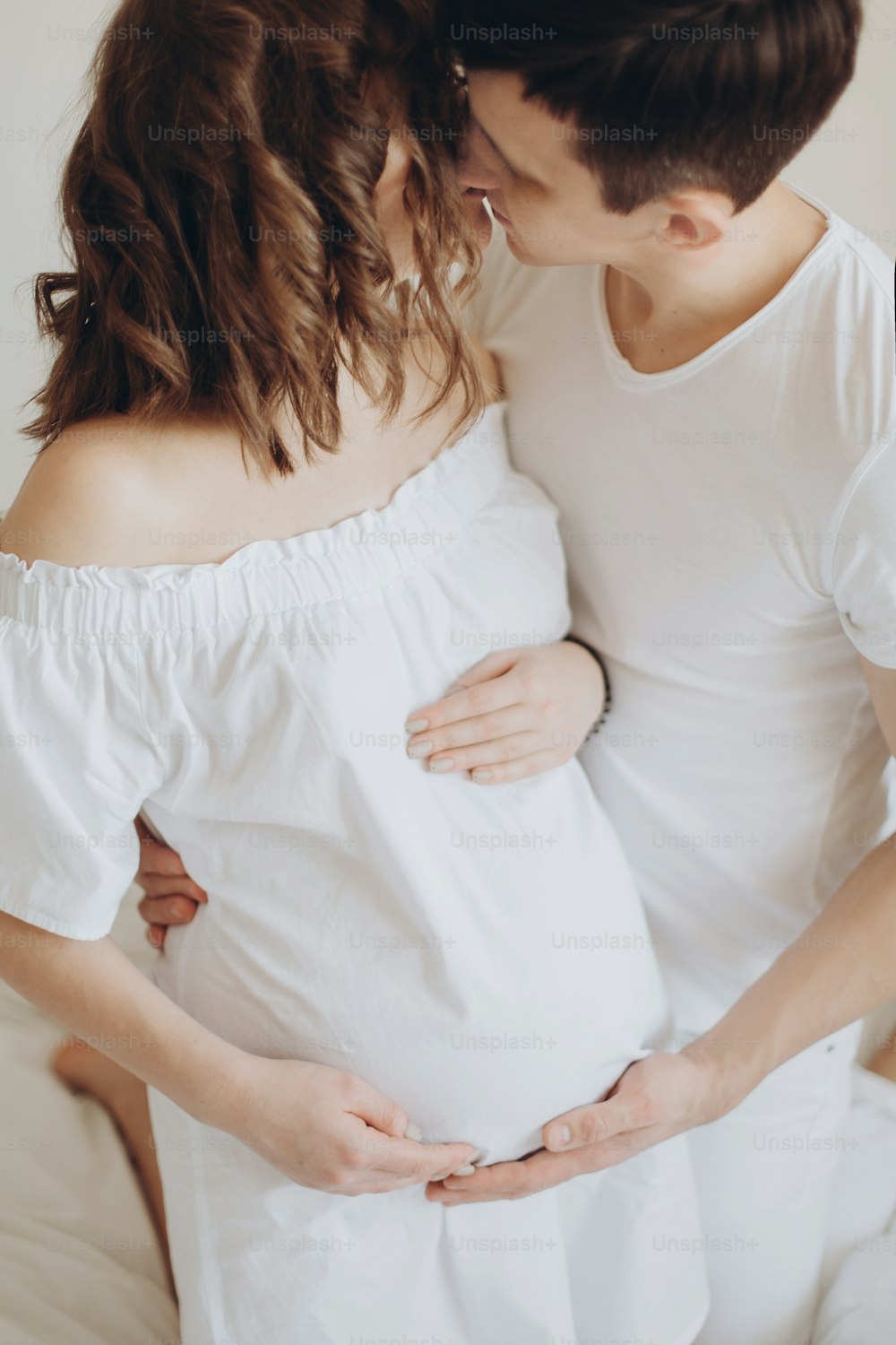 Glückliches schwangeres Paar, das sich auf einem weißen Bett entspannt und Bauchstoß hält. Glücklicher junger Ehemann, der seine lächelnde Frau küsst und den Babybauch umarmt.  Stilvolle schwangere Familie in Weiß zu Hause. Wahres Glück