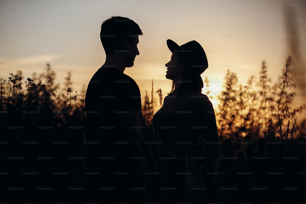 Stylish pregnant couple silhouettes in sunny light in autumn park among grass. Happy young parents, mom and dad, hugging baby bump, enjoying beautiful moment at sunset. Parenthood concept