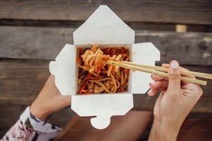 Girl eating wok noodles with vegetables and seafood in carton box to go, with bamboo chopsticks, closeup. Traditional Asian cuisine at Street food festival. Thai noodles in paper box takeaway