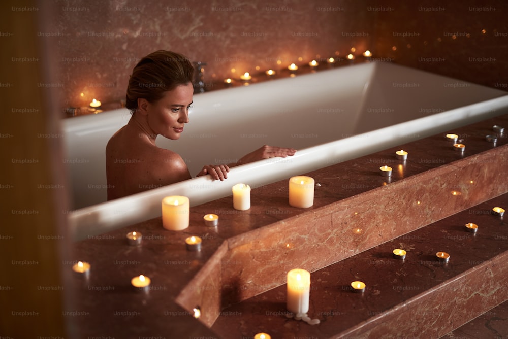 Concept of relaxation and body care. Top angle portrait of calmly smiling woman taking spa bath with lighten candles around