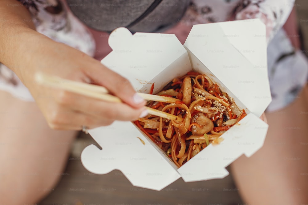 Wok with noodles and vegetables in carton box to go and bamboo chopsticks. Traditional Asian cuisine. Asian Street food festival. Girl eating thai noodles in open box takeaway. Food delivery