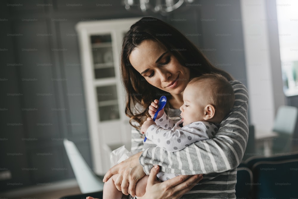 Beautiful woman holding her child in arms and admiring him. He is keeping a spoon in his small hands. First kid's food concept