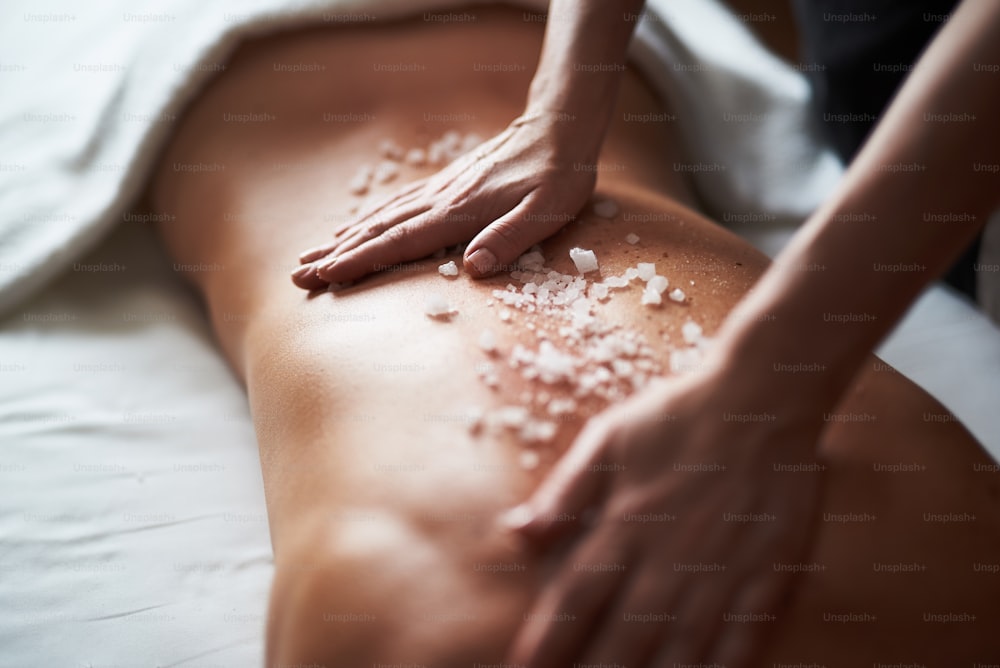Concept of relaxation and body care. Close up portrait of therapist applying mineral salt on back of young lady while making healthy massage in spa beauty salon