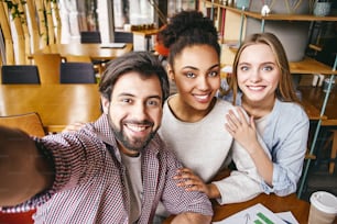 Three laughing young business people in casual wear smile to make shot of themselves. Office interior, bookshelves in the background. Concept of success