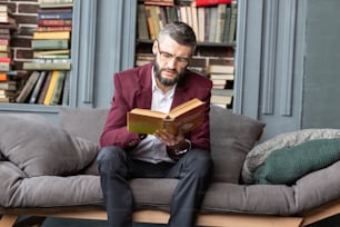 Reading book. Bearded businessman wearing dark red jacket feeling involved in reading book about money