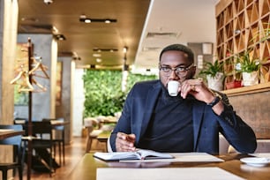 Gros plan d’un jeune PDG afro-américain élégant en veste bleu foncé en train de parcourir de la paperasse, assis à la table d’un café avec des documents et prenant des notes dans un carnet tout en buvant le café du matin dans un café