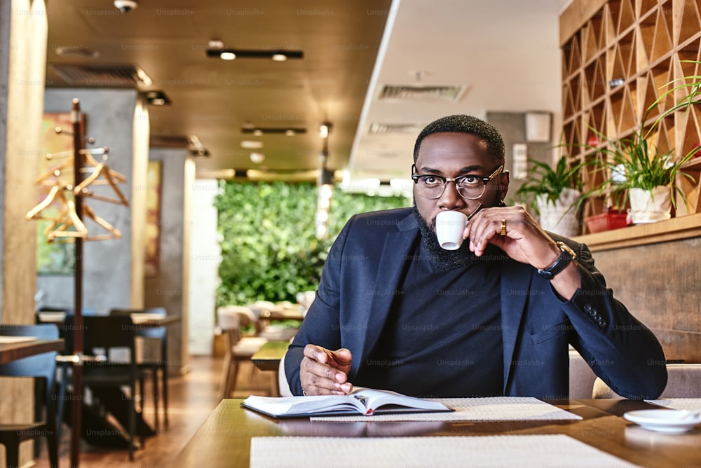 Close up of stylish young African-American CEO in dark-blue jacket going through some paperwork, sitting at cafe table with documents and making notes in notebook while drinking morning coffee in cafe