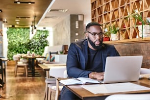 Portrait of dark-skinned businessman in dark-blue jacket and glasses sitting at the table, indoors, making notes, working with his laptop computer. Young African entrepreneur is coming up with new ideas. Modern interior
