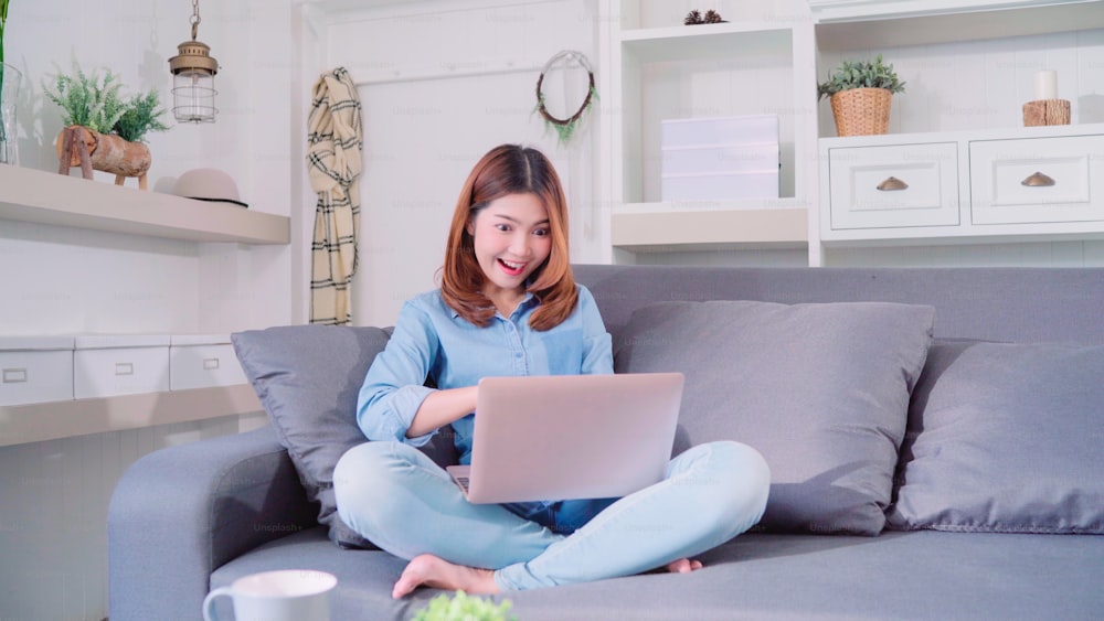 Ritratto di bella giovane donna asiatica sorridente attraente che utilizza computer o laptop mentre è sdraiata sul divano quando si rilassa nel soggiorno di casa. Godersi il tempo stile di vita delle donne a casa concetto.