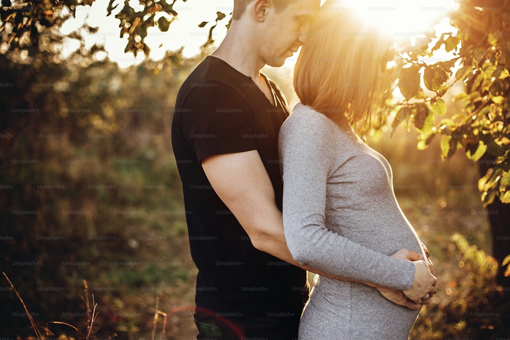 Happy young parents, mom and dad, hugging baby bump, enjoying beautiful moment at sunset. Stylish pregnant couple holding hands on belly and embracing in sunny light in autumn park under tree