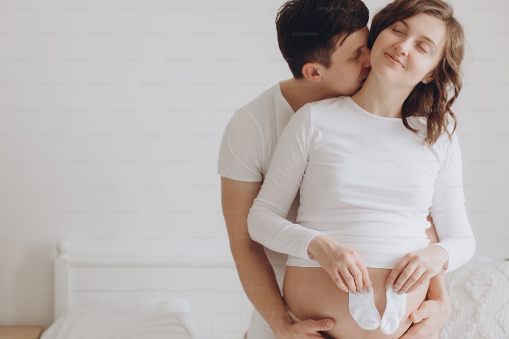 Happy young pregnant couple holding white little socks on belly bump and relaxing on white bed. Stylish pregnant family, mom and dad in white, kissing at home and hugging belly. True happiness