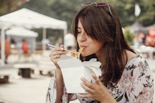 Stylish hipster girl eating wok noodles with vegetables from carton box with bamboo chopsticks. Asian Street food festival. Happy boho woman eating noodles in takeaway paper box