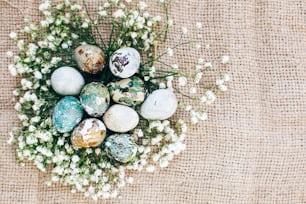 Stylish Easter quail eggs with spring flowers in floral nest on rustic fabric in sunny light on wood. Modern colorful eggs painted with natural dye in blue, green. Happy Easter, greeting card