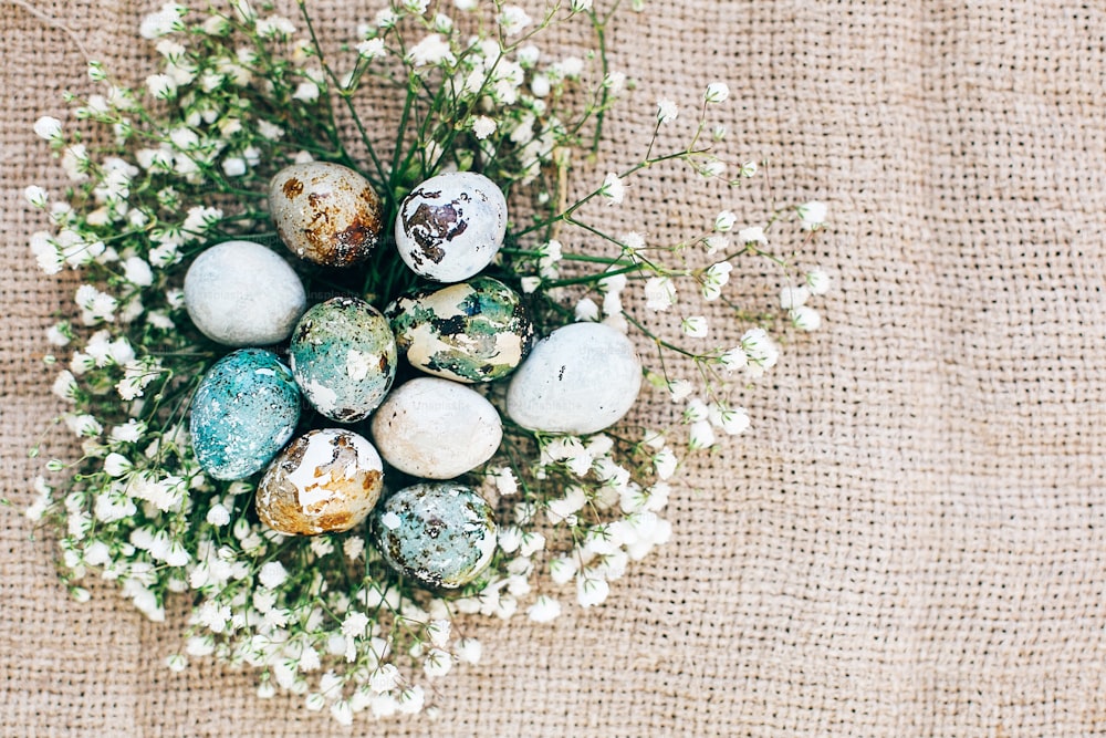 Stylish Easter quail eggs with spring flowers in floral nest on rustic fabric in sunny light on wood. Modern colorful eggs painted with natural dye in blue, green. Happy Easter, greeting card