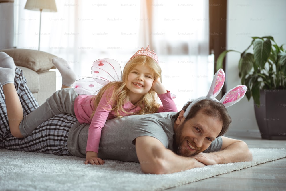 Lustiges Spiel. Porträt eines fröhlichen Mannes liegt auf dem Boden, während er sein Kind auf dem Rücken hält. Er schaut in die Kamera und lächelt. Mädchen posiert mit Feenflügeln und rosa Krone