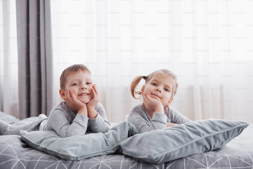 Happy kids playing in white bedroom. Little boy and girl, brother and sister play on the bed wearing pajamas. Nightwear and bedding for baby and toddler. Family at home.