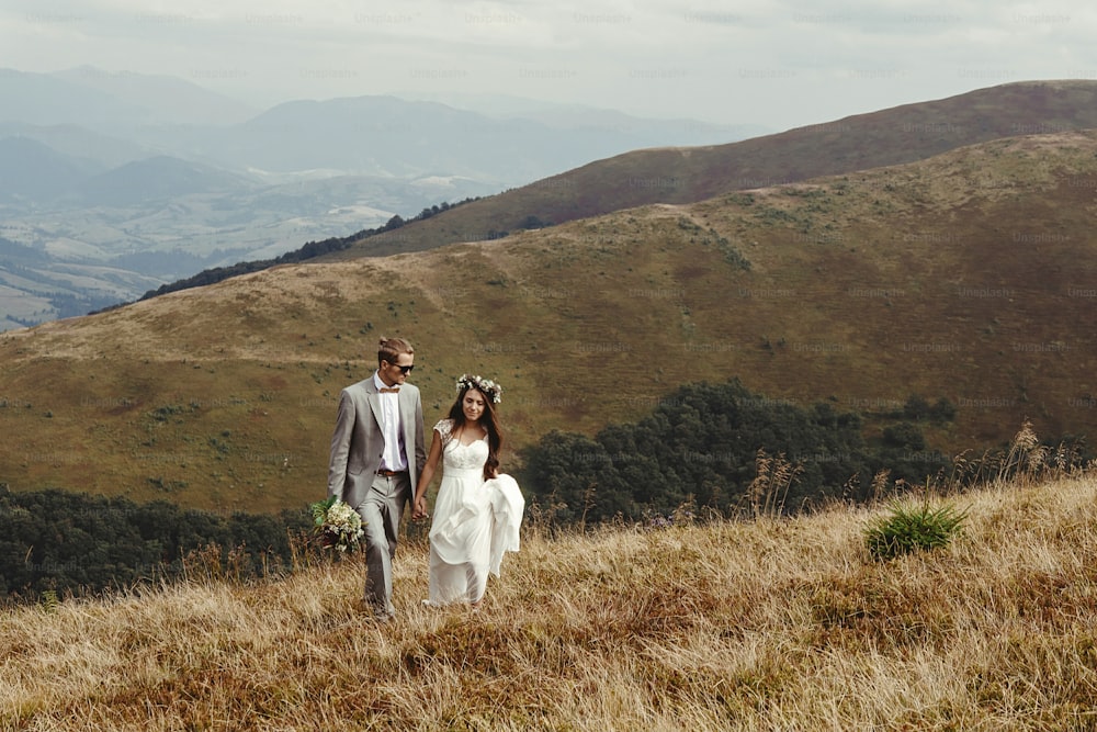 happy gorgeous bride and groom walking  in sun light holding hands, boho wedding couple, luxury ceremony at mountains with amazing view, space for text