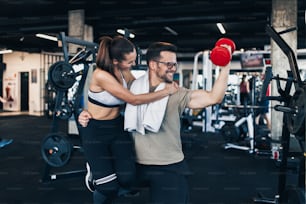 Young attractive couple working out smiling and having fun in modern fitness gym. Man holding woman with one hand and red dumbbell with another.