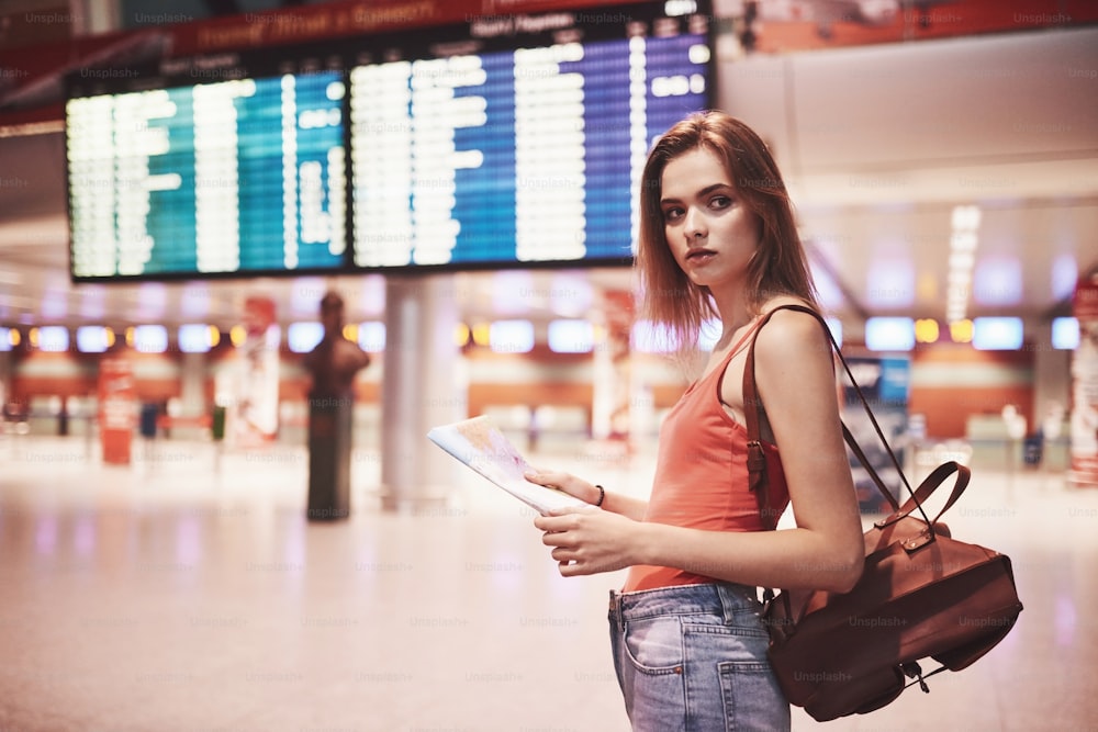 Beautiful young tourist girl with backpack in international airport, near flight information board.