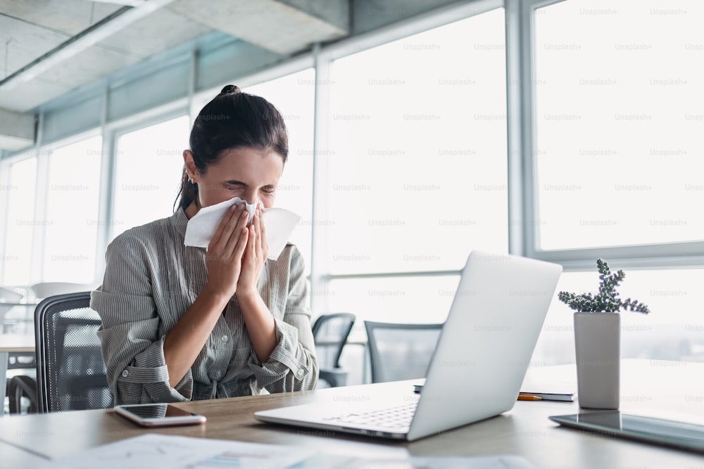 Ill young dark-haired woman caught cold sneezing in tissue in office, sick allergic girl blowing, wiping running nose, got flu, rhinitis, sinusitis, coughing, having seasonal allergy symptoms at workplace