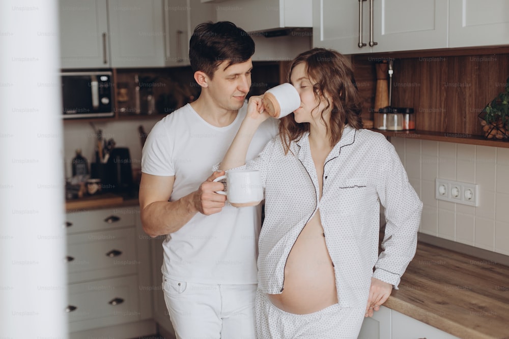 Happy young pregnant couple in white pajamas holding cups and relaxing in kitchen in the morning. Stylish pregnant family, mom and dad at home, waiting for baby. Cute moments of happiness.