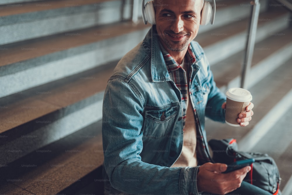 Photo of attractive and friendly adult man sitting on stairs with beverage in hand. He listening music in on cellular wearing headset