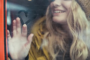 Sending love. Close up low face portrait of hipster cheerful lady inside of transport drawing heart shape on steamy window