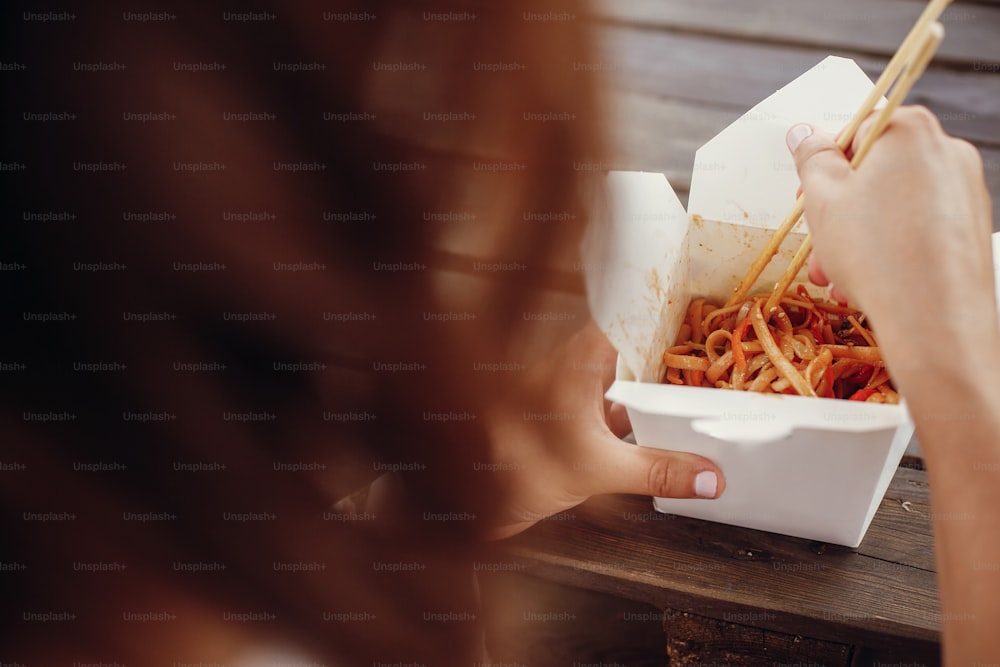 Girl eating wok noodles with vegetables and seafood in carton box to go, with bamboo chopsticks, closeup. Traditional Asian cuisine food delivery. Noodles in paper box takeaway. Street food