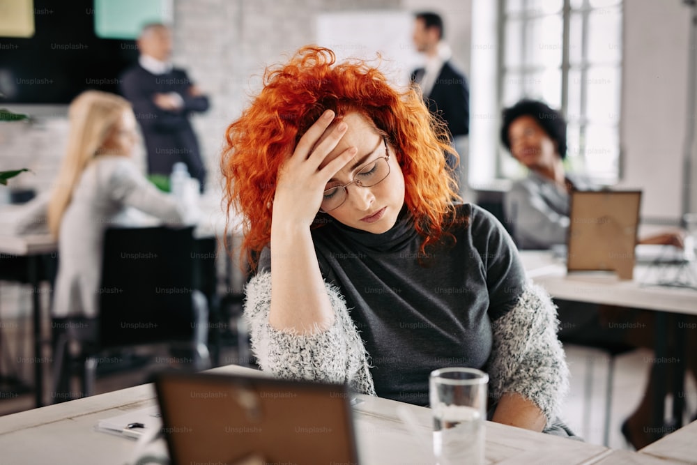 Empresária deprimida segurando a cabeça com dor e se sentindo deprimida no trabalho. Há pessoas em segundo plano.