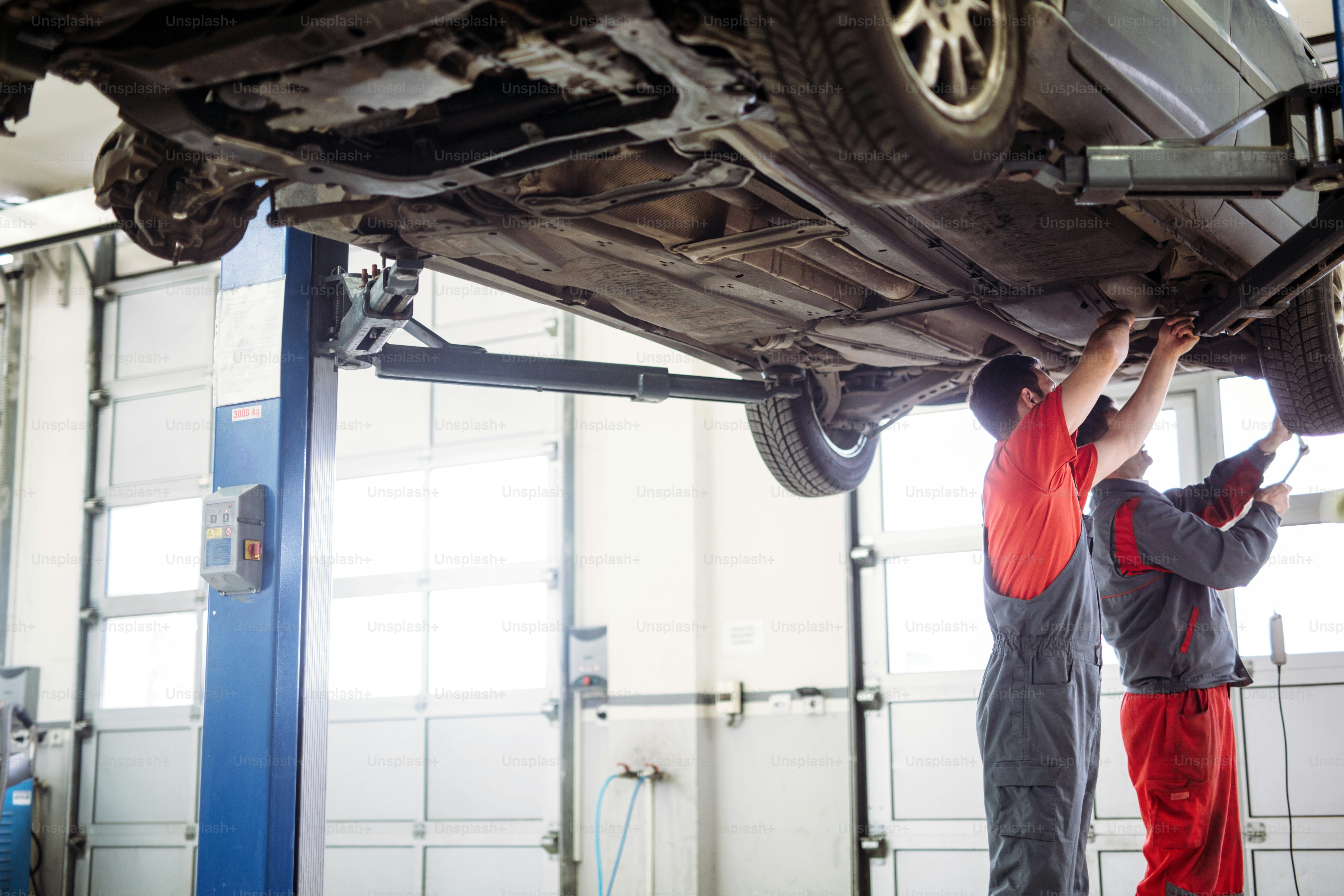 Two handsome mechanics in uniform are working in auto service with lifted vehicle.