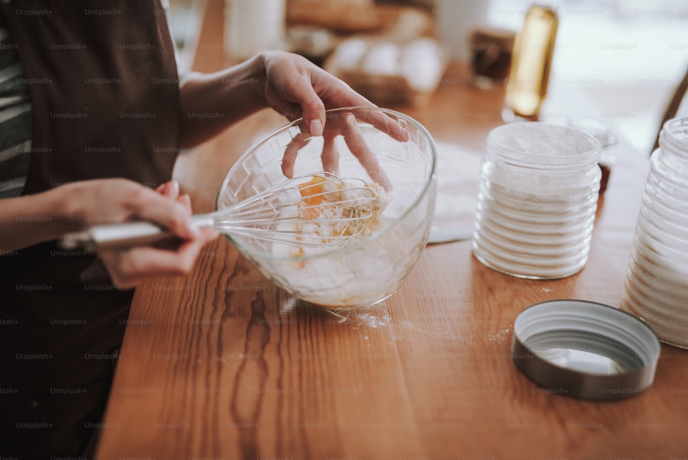 Top view of female mixing flour with eggs while using whisk. She is cooking bakery in domestic atmosphere