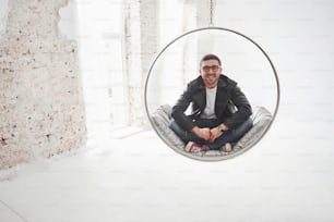 Full-length portrait of relaxed hipster man in casual sitting on hanging chair bullet and smiling on camera isolated over white background.