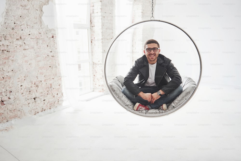 Retrato de corpo inteiro de um homem hipster relaxado em casual sentado na bala da cadeira pendurada e sorrindo na câmera isolado sobre o fundo branco.