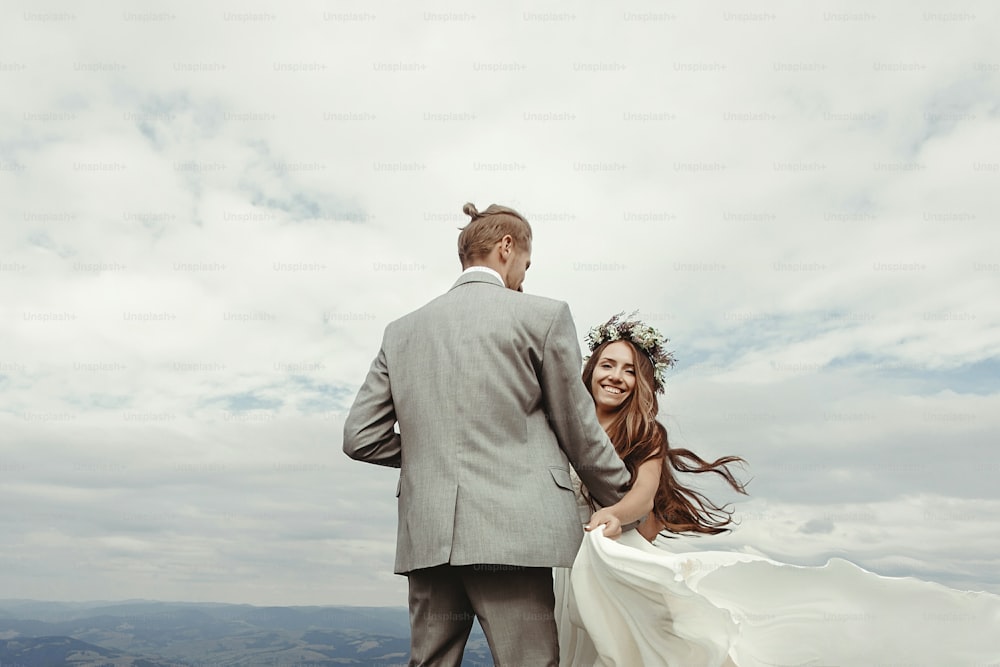 gorgeous bride and groom holding hands and dancing at sky and clouds, moment of true happiness, luxury ceremony at mountains with amazing view, space for text