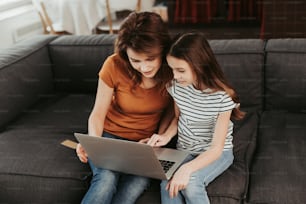 Close family relationship. Waist up top angle portrait of happy smiling woman and her female child looking in laptop while sitting on sofa at home