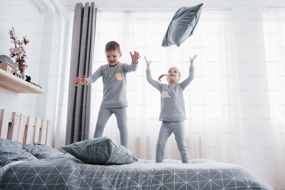 Happy kids playing in white bedroom. Little boy and girl, brother and sister play on the bed wearing pajamas. Nightwear and bedding for baby and toddler. Family at home.
