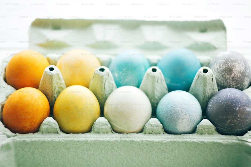 Stylish Easter eggs in carton tray on white wooden background, space for text. Modern colorful easter eggs painted with pastel natural dye in rainbow colors. Happy Easter. Holiday decor